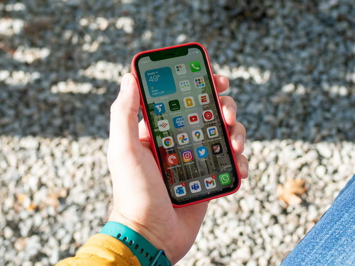 A hand holds an Apple iPhone 12 mini with the home screen showing against a gravel backdrop.